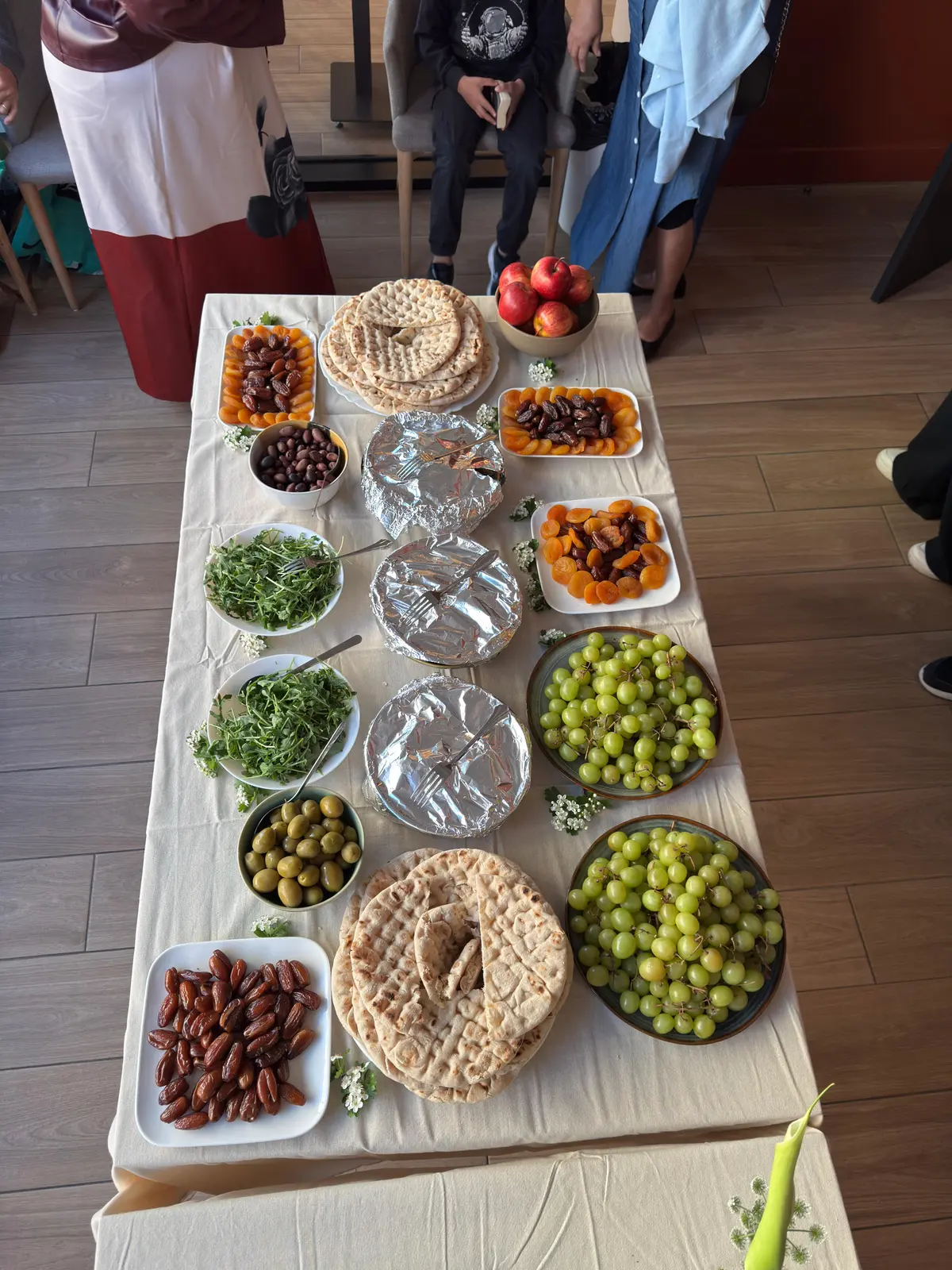 Mesa comprida de buffet com pão pita, frutos secos, uvas, azeitonas e salada.